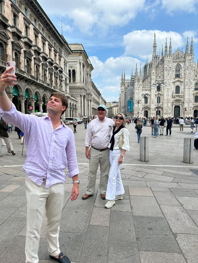 Three people taking a selfie in a public square with the Milan Cathedral in the background under a blue sky with clouds.